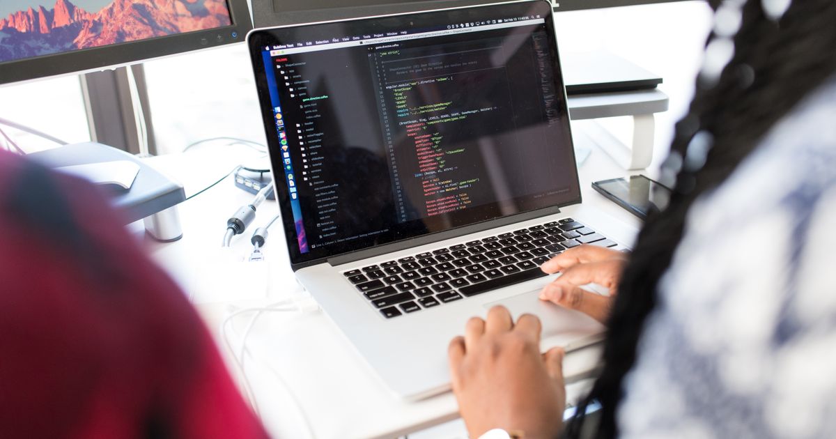 Over the shoulder view of a person working at a laptop with a code editor open on screen, a second monitor beside it on a bright desk.