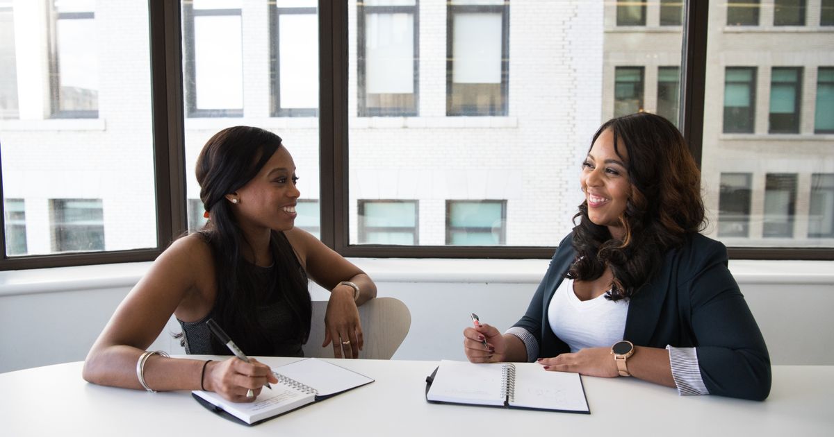 Two colleagues sitting at a white office table by a window, open notebooks and pens in front of them, going through figures together.