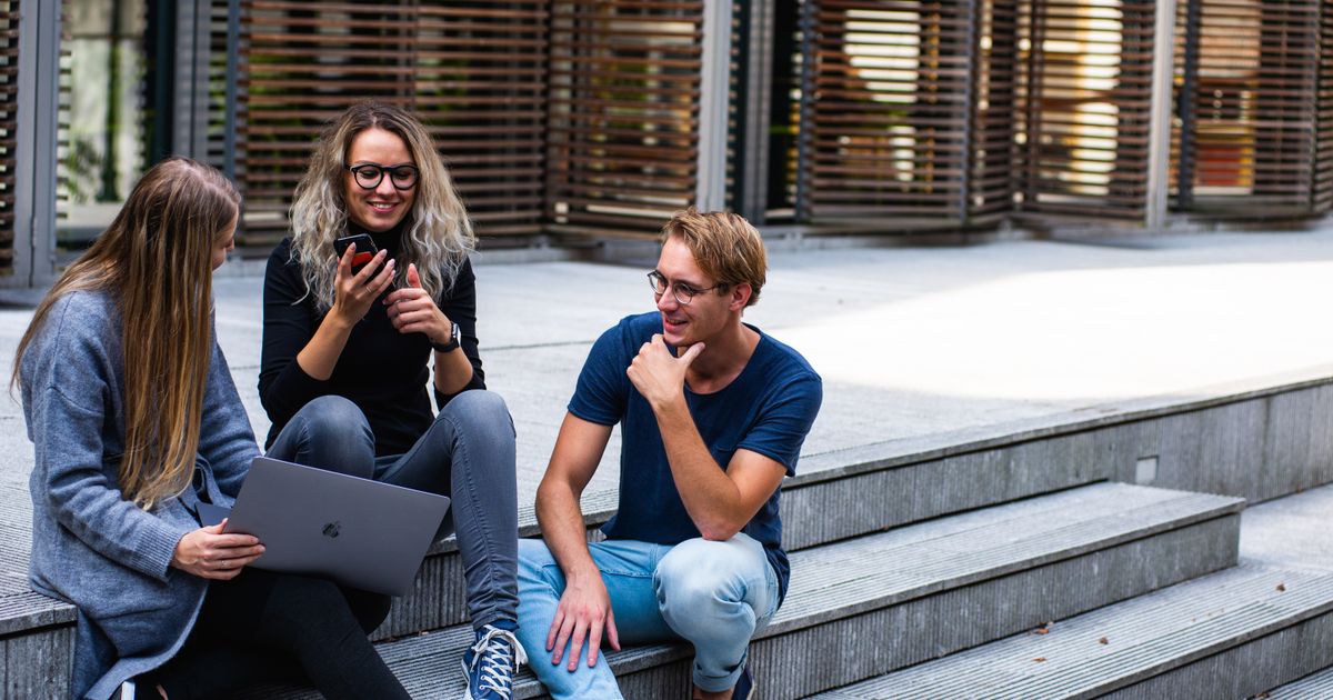 Three colleagues sitting on outdoor steps comparing prospecting tools on a laptop and a phone